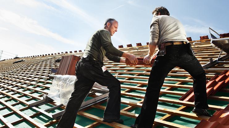 Bild fehlt Zwei Handwerker arbeiten auf einem geneigten Dach, während sie Holzlatten tragen und das Dach decken.