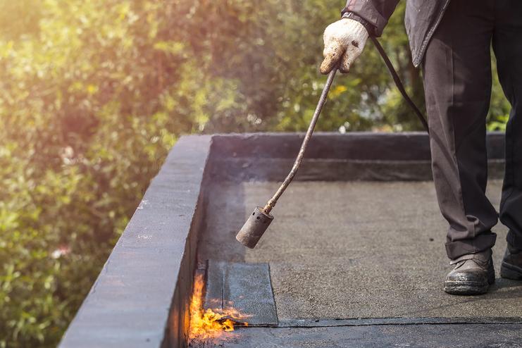 Handwerker mit Lötbrenner schweißt Bitumenbahnen auf einer Dachoberfläche, Flamme sichtbar.