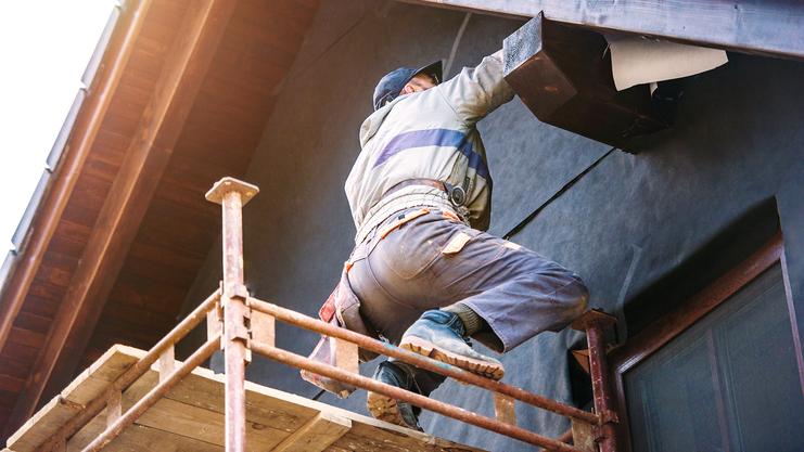 Arbeiter steht auf einem Gerüst und befestigt ein Element unter dem Dach eines Hauses.